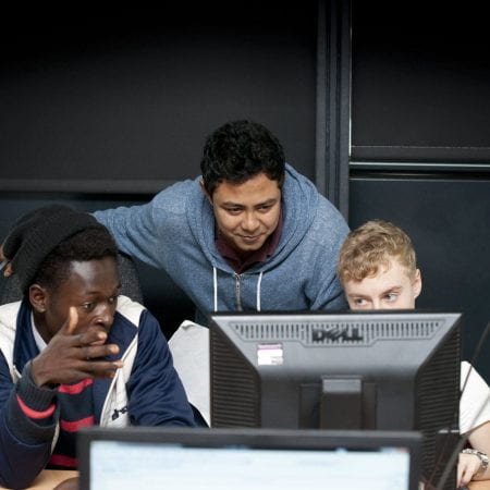 An image of three young students talking and sat in front of a black computer. .